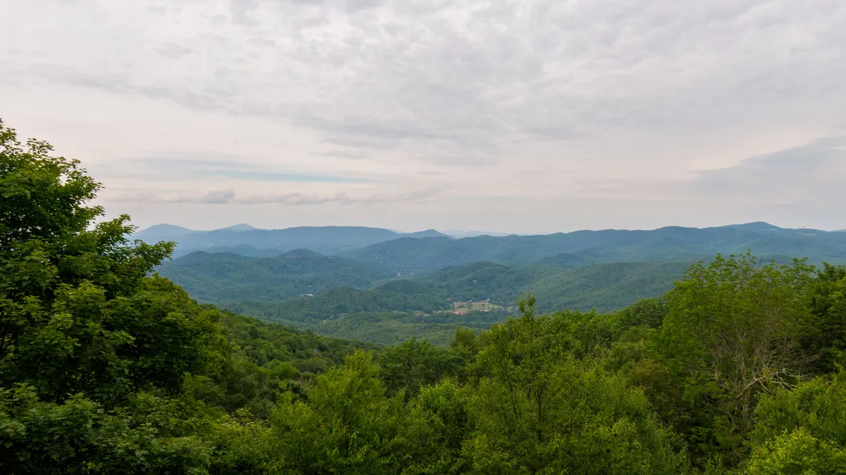 A sweeping view of the Blue Ridge mountain range framed by lush green trees under a picturesque cloudy sky. The tranquil scene captures the grandeur of the natural setting and hints at the stunning sunsets and outdoor experiences available to guests.