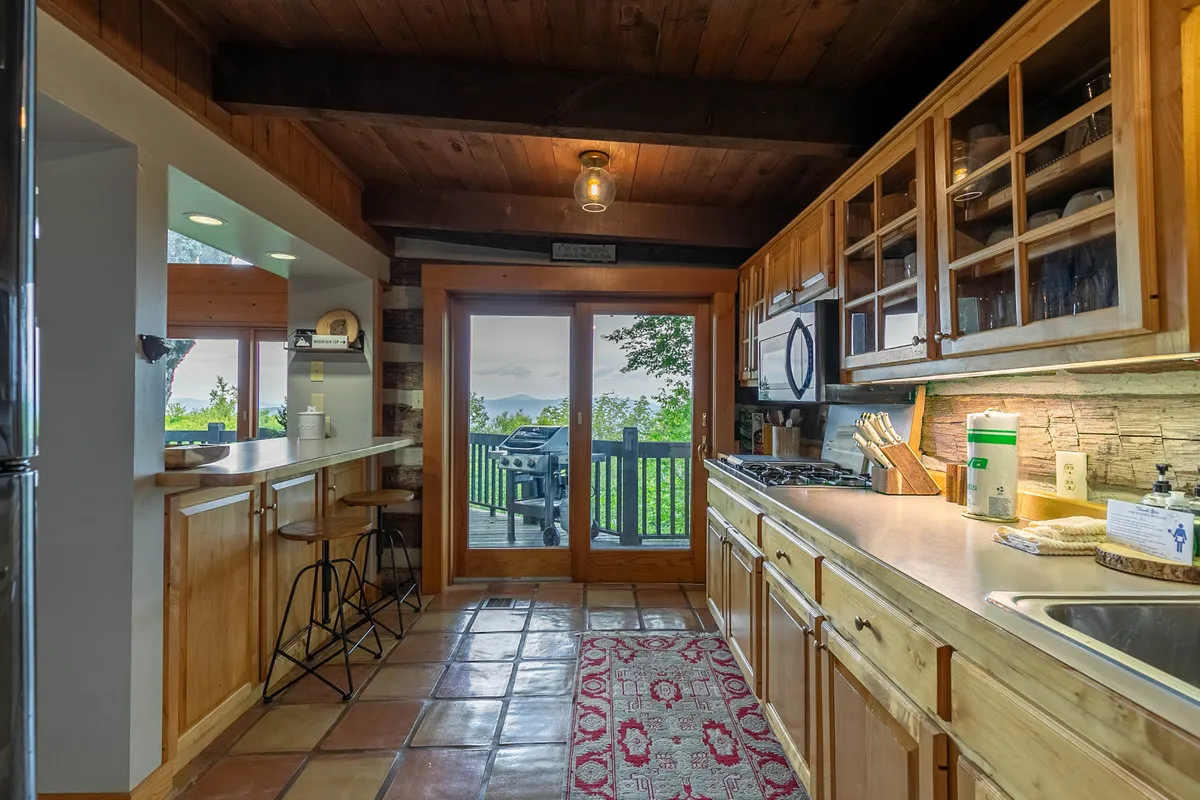 A spacious kitchen with large windows that frame expansive mountain views. Wooden cabinetry and a kitchen island create a warm, inviting atmosphere, and bar seating faces directly toward the scenery outside. The space is filled with natural light, suggesting a seamless connection to the outdoors.