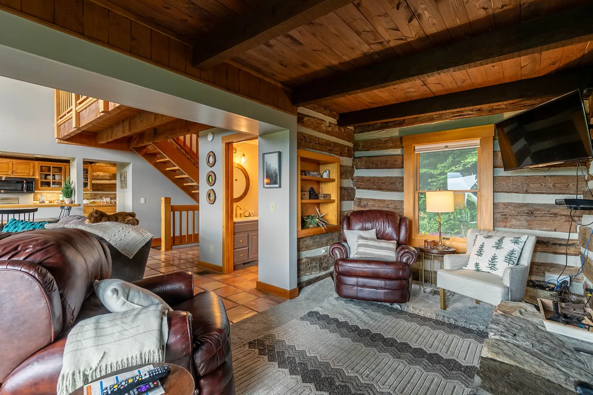 This image features a welcoming lounge space with leather armchairs, wooden beams, shelves with decorative items, and a large window letting in natural light. The setting is ideal for relaxing with your pet after time spent outdoors, and the visible window maintains a visual link to nature.