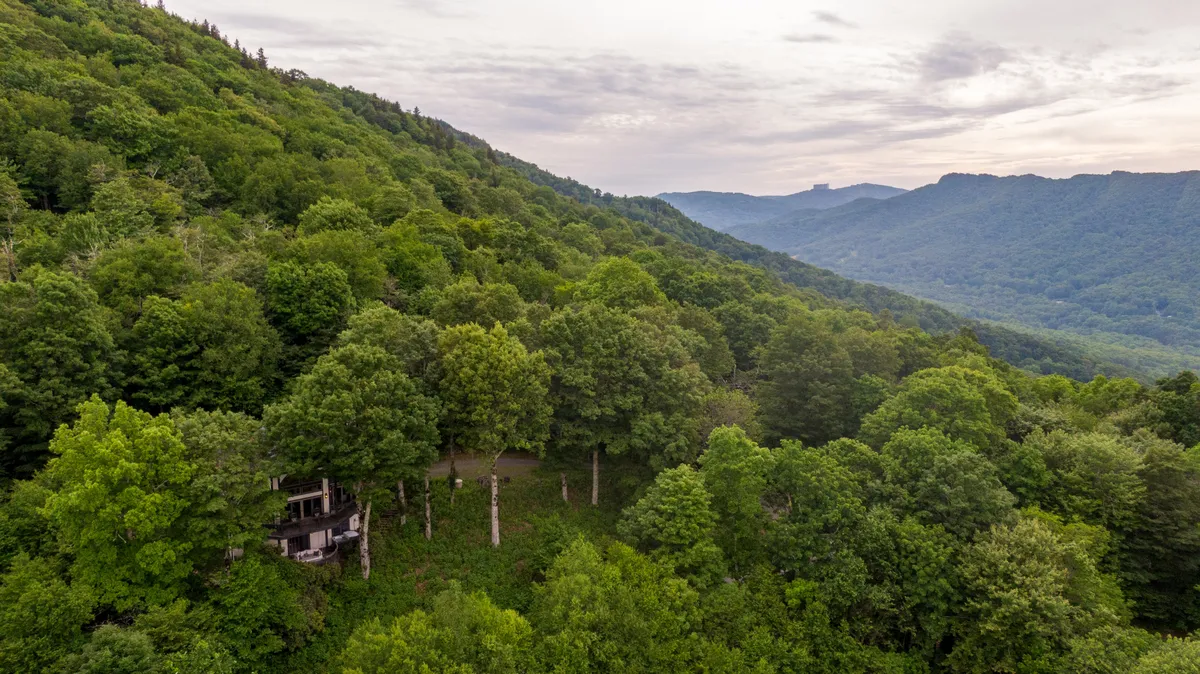 This image shows a modern house nestled among lush green trees with a stunning mountain range in the background. The scene is peaceful and invites viewers to imagine stepping right out into hiking trails, nature walks, and bird watching. The expansive mountain views evoke a sense of adventure and exploration, perfectly matching the High Country setting.