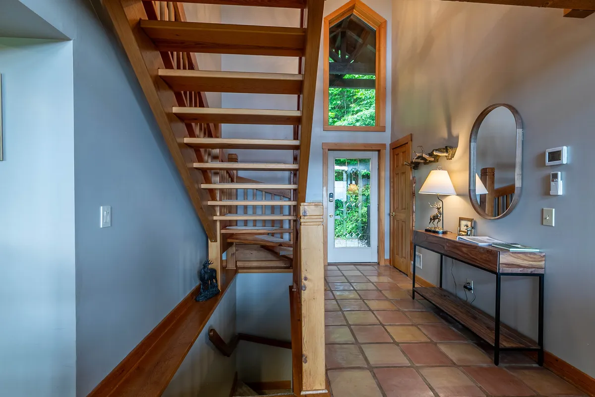 A lofted area with a staircase leading to a cozy landing featuring a large window. The console table and lamp suggest a flexible spot for work or relaxing, while the window visually connects interior spaces to the forested outdoors.