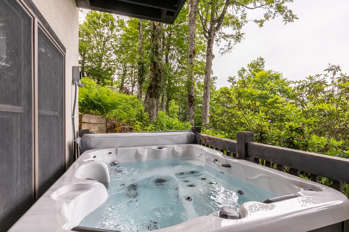 A bubbling hot tub sits on a wooden deck, surrounded by greenery and enclosed by a sturdy railing. The hot tub is positioned to maximize views of the natural landscape, offering a cozy spot for relaxation after a day in the mountains.