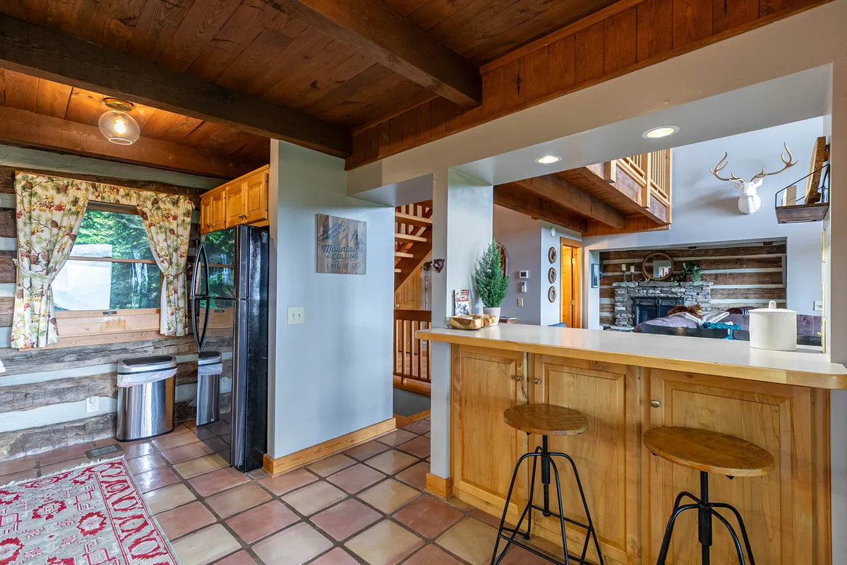 An open-concept kitchen and living area featuring a kitchen island, large window with floral curtains, stainless steel appliances, exposed wooden beams, and a stone fireplace. The area is styled for both meal preparation and relaxation by the fireplace, fostering togetherness.