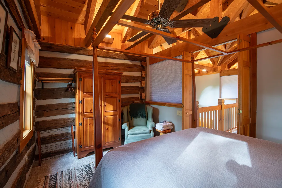 A warm, inviting bedroom featuring a four-poster bed, wooden armoire, and exposed beams. A green armchair is positioned by a window, offering a quiet nook to read, relax, or contemplate the views beyond.