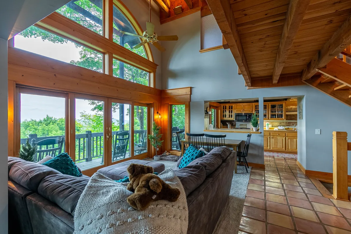 This image shows a bright, open living area with large windows, a comfortable sofa, a wooden staircase, and a visible kitchen area. The expansive windows connect the interior with the outdoors, suggesting easy access to nature and providing scenic views, which are ideal after a day of exploring the trails with pets.