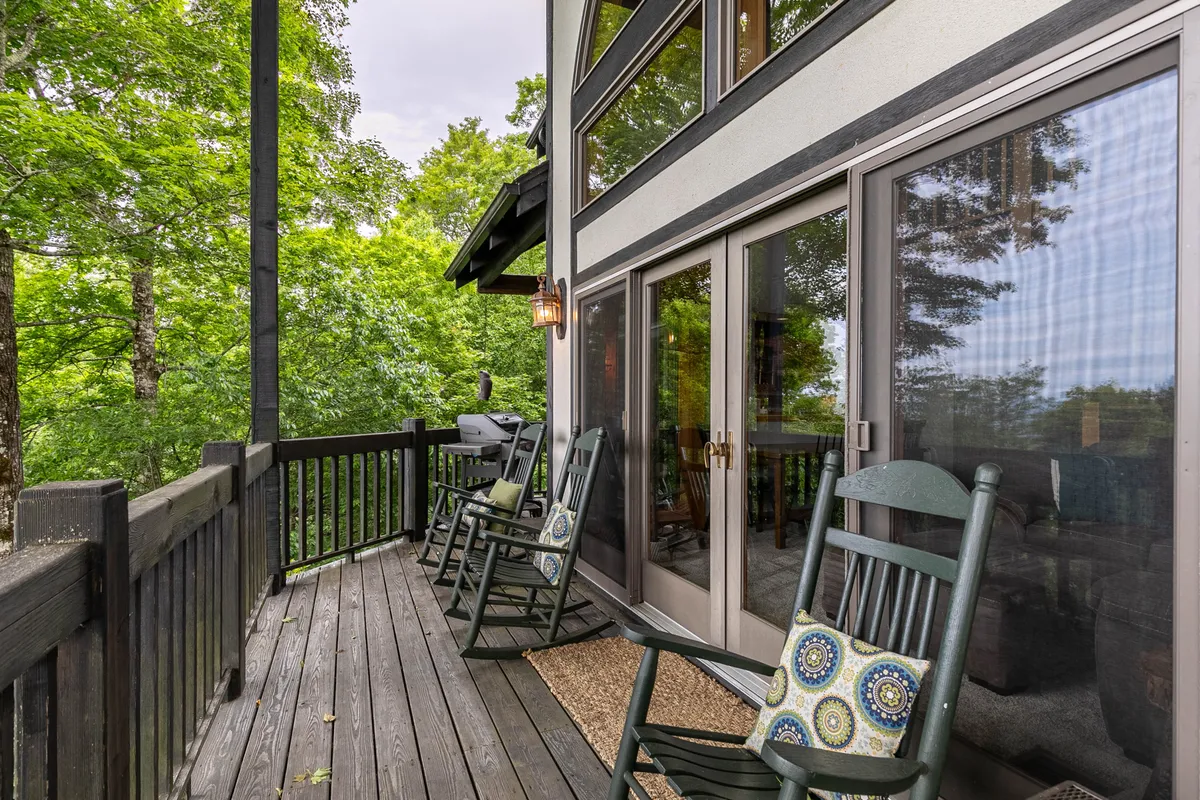 This exterior deck scene features wooden rocking chairs set against large windows, surrounded by green trees. The inviting space suggests a seamless indoor-outdoor connection and a calming spot for enjoying nature or socializing.