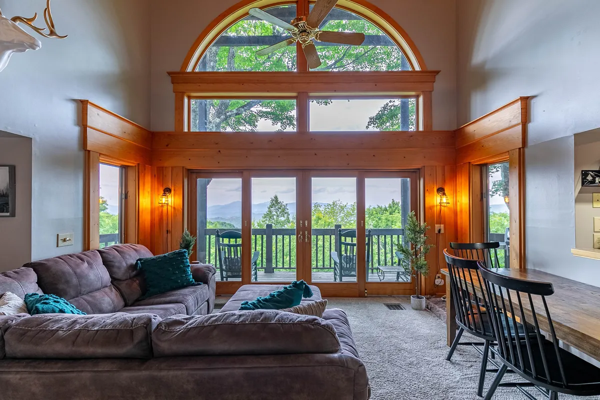 A spacious living area with large windows framing sweeping mountain views. A comfortable couch sits near the window, with a dining area and wooden accents contributing to a cozy, inviting atmosphere. Soft natural light pours in, making the space feel bright and connected to the outdoors.