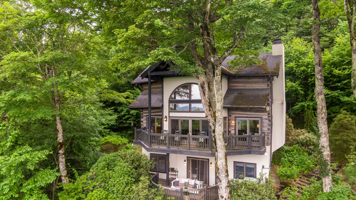 The image showcases the exterior of a rustic chinked log cabin nestled among lush green trees, with a spacious deck overlooking breathtaking mountain vistas. The setting is serene, inviting guests to relax on the deck while soaking in panoramic sunrise views and the beauty of surrounding nature.