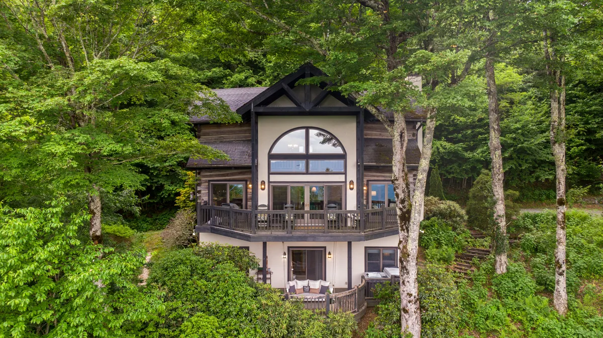 The image displays a home with large windows and a balcony, surrounded by trees. The outdoor setting hints at the possibility of storytelling by the fire pit, enjoying sunsets, and connecting with the natural environment from the comfort of the cabin’s exterior spaces.