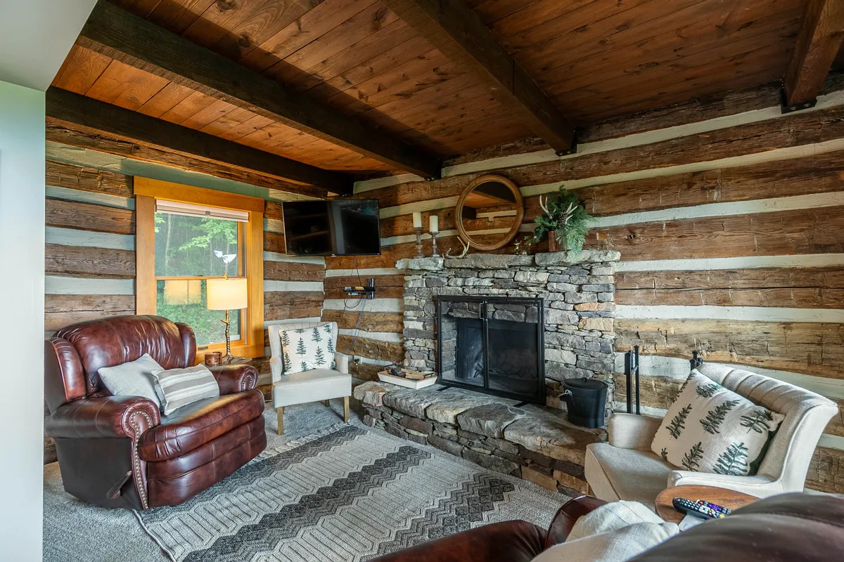 A warm and inviting den area with a stone fireplace, leather armchair, and rustic wooden beams overhead. The space evokes feelings of comfort, perfect for curling up with a book or gathering for games and intimate conversation.
